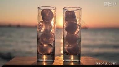 两个玻璃杯的时间间隔特写镜头与融化的冰在大海和日落的背景